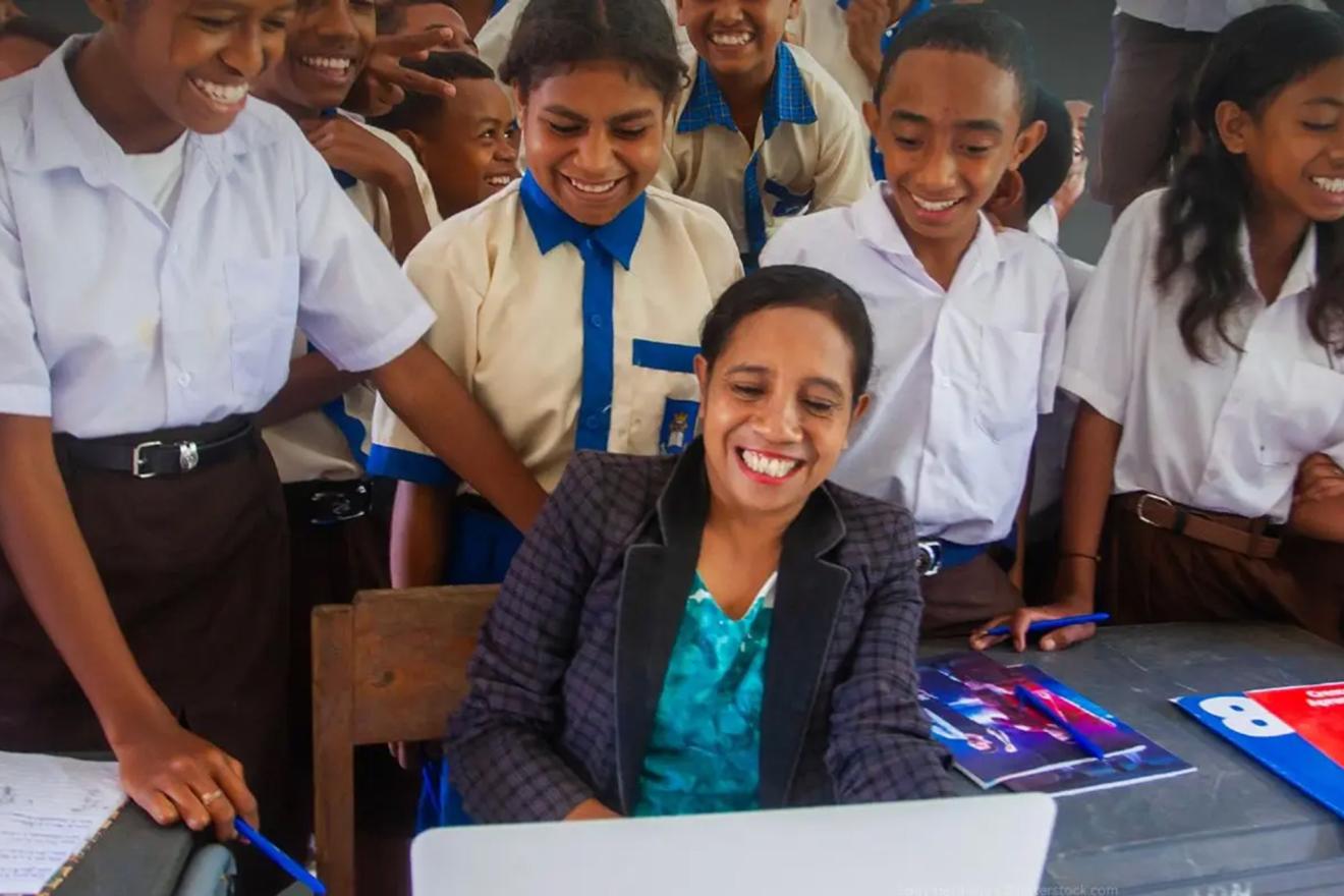 A classroom scene where a group of students gather around a teacher seated at a desk with a laptop.
