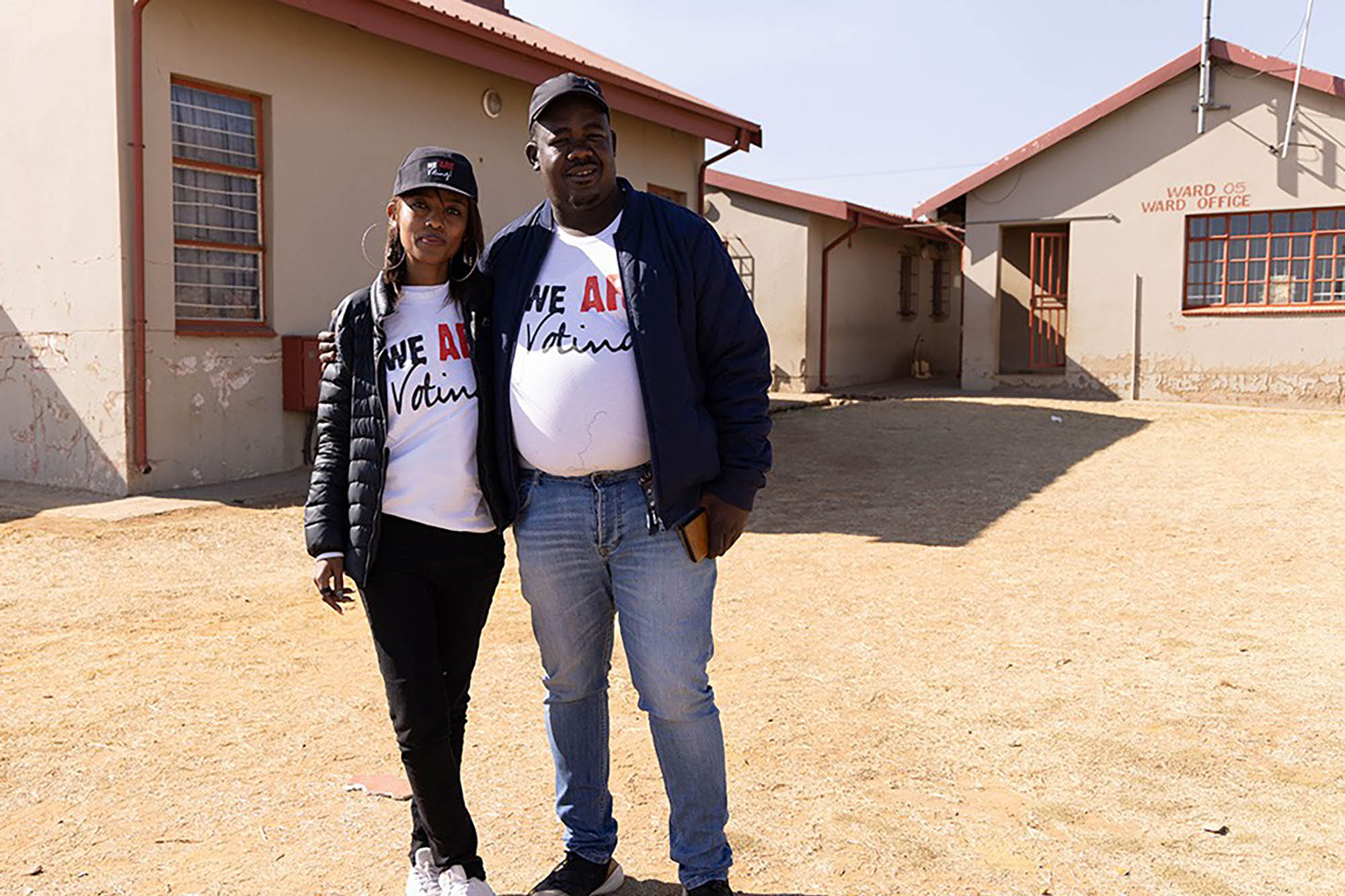 Two individuals standing outside a building wearing white T-shirts that say, "WE ARE Voting".