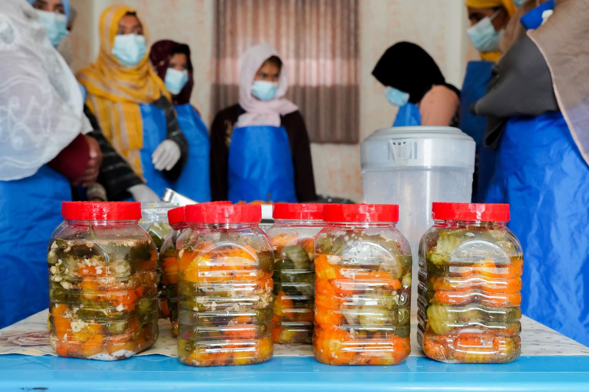 A group of women wearing blue aprons gathering around a table where there are several jars of pickled vegetables, including carrots and green peppers arranged in two rows.