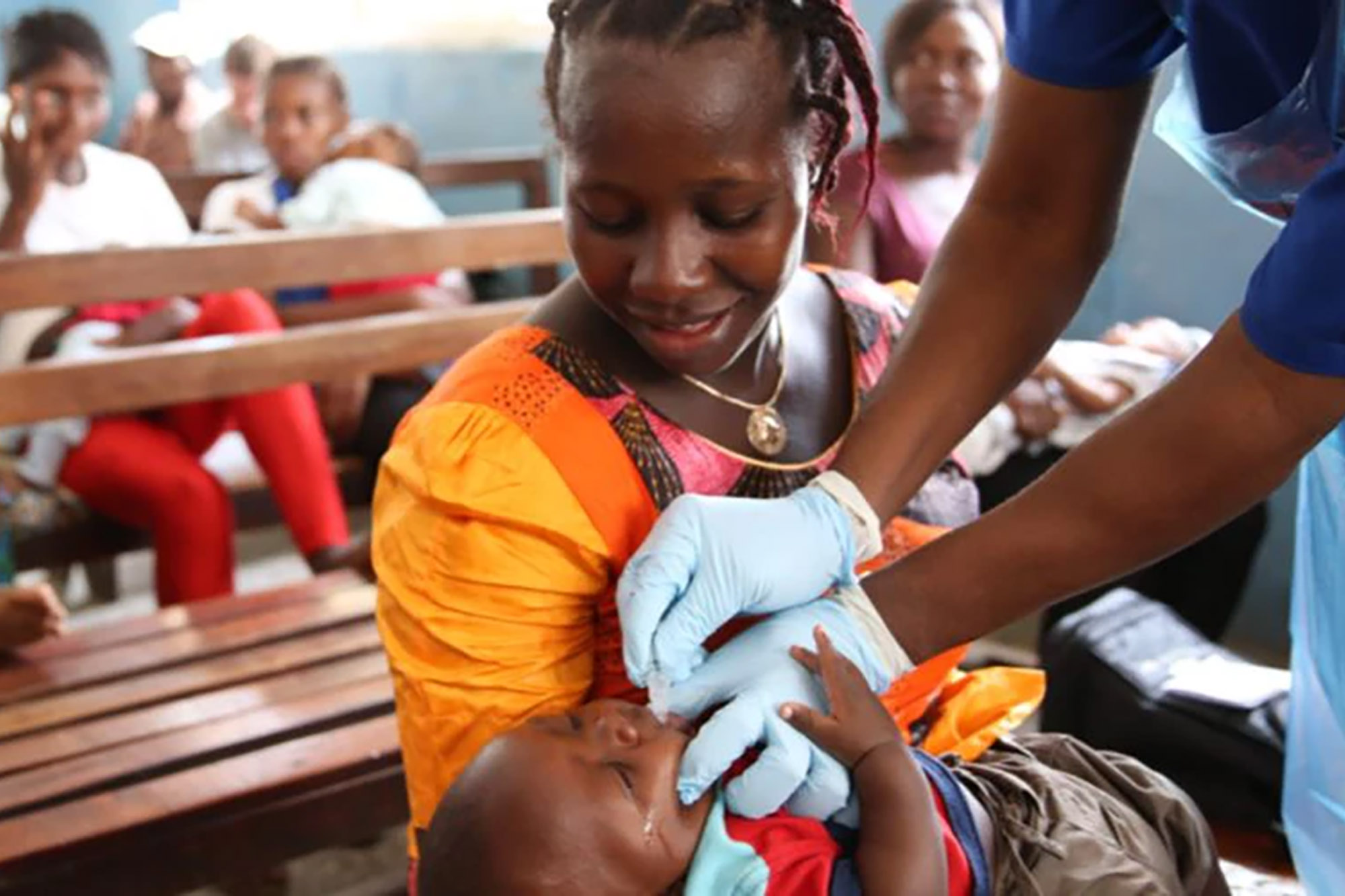 A vaccination center with a medical professional in a blue administering an injection to a child. The child is being held by an adult dressed in an orange and yellow garment. 