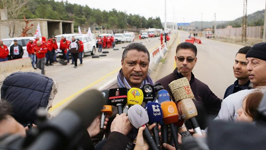 Yacoub stands in the middle of a highway. Many journalist microphones at his face and a group of people in the distance behind wearing red jackets