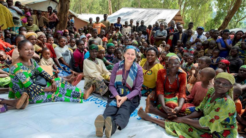 Group photo with a large assembly of women, children and men outdoors surrounded by trees and fragile homes some seated on a UNICEF banner