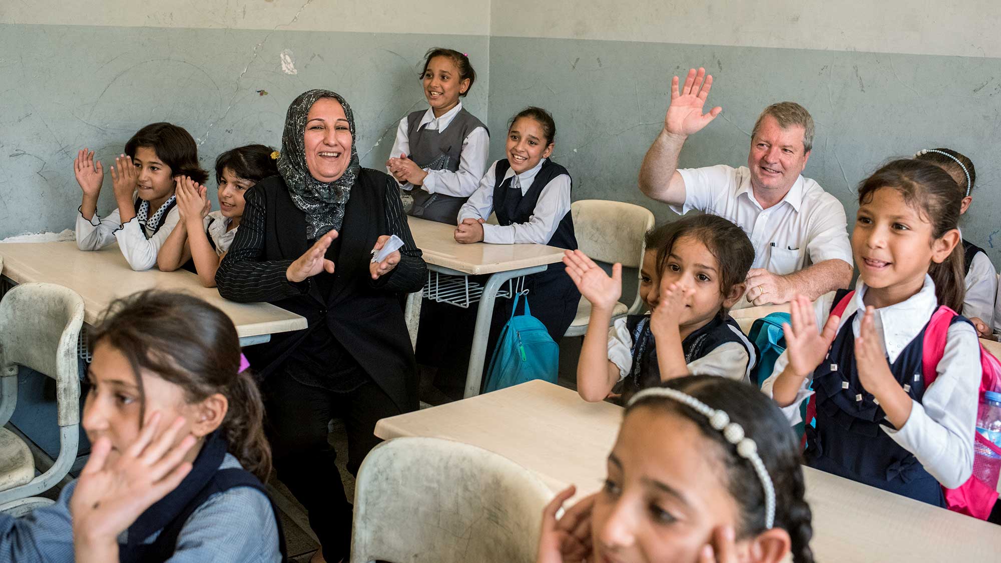 Peter is raising his hand whilst sitting in a classroom with children and a teacher all of whom are clapping their hands