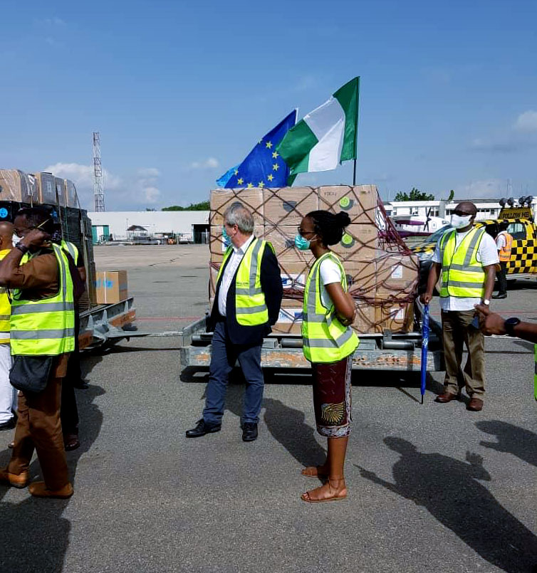 Peter stands in the outdoors among planes, people, boxes and two flags one of which is the EU flag