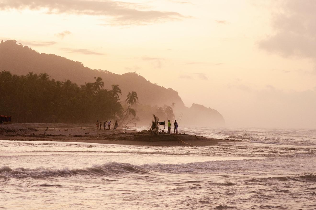 Paisaje costero al atardecer con mar agitada y varias personas pescando de fondo. 